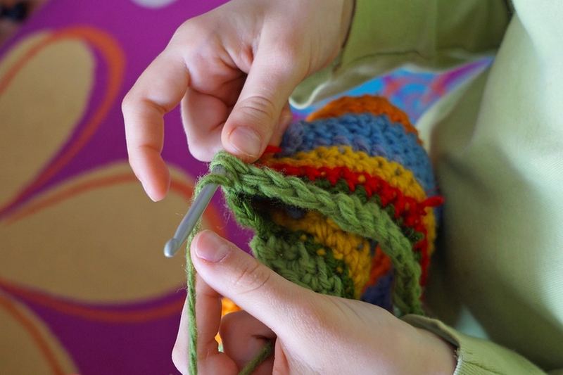 Manos tejiendo a crochet con aguja verde sobre una labor de rayas de colores
